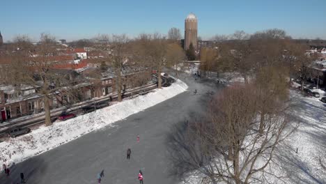 Sideways-aerial-pan-revealing-urban-winter-scene-with-people-ice-skating-along-a-curved-frozen-canal-going-through-the-Dutch-city-of-Zutphen-with-former-water-tower-in-the-background