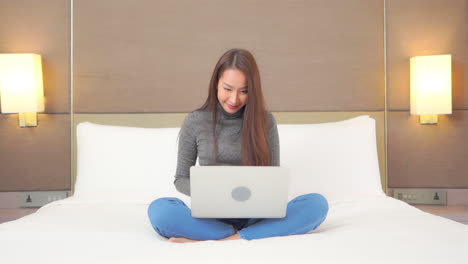 A-young-woman-comfortably-works-on-her-laptop-while-sitting-on-a-huge-hotel-suite-bed
