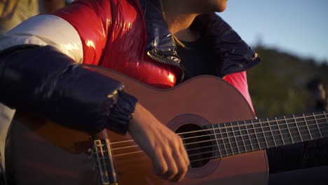 Young-female-with-shiny-jacket-playing-guitar-at-sunset-outdoors-in-mountains-slow-motion-60fps