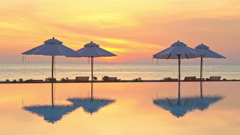 A-colorful-ocean-sunset-reflects-its-vibrant-color-and-beach-umbrellas-into-a-resort-infinity-edge-swimming-pool