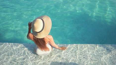 A-woman-with-her-back-to-the-camera-while-sitting-in-the-shallow-water-of-a-pool-step-holds-the-edge-of-her-sunhat-as-she-looks-around