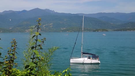Kleines-Segelboot-Einsam-In-Einem-See-Mit-Bergen-Und-Wald-Im-Hintergrund-Und-Grünen-Blättern-Vor