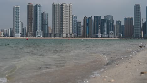 Sharjah:-Meerwasser-Trifft-Und-Bricht-An-Einem-Strand,-Meereswellen-Und-Städtischer-Wolkenkratzer-Der-Stadt-Sharjah-Im-Hintergrund