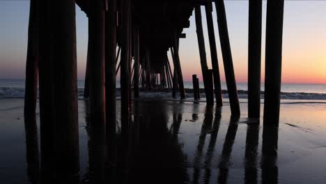 Slow-motion-sunrise-view-of-waves-rolling-in-and-crashing-against-the-wooden-supports-of-a-fishing-pier-on-a-beach