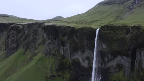 Powerful-cascade-flows-off-steep-cliff-in-Iceland-during-cloudy-day,-aerial