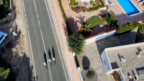 Artistic-birdseye-drone-footage-of-three-team-cyclists-riding-along-a-straight-road-in-Calpe,-Spain,-on-a-warm-morning-with-long-shadows,-and-traditional-terracotta-roofed-buildings-lining-the-road