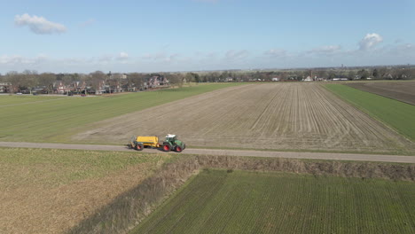 Aerial-of-tractor-with-large-tank-driving-between-green-meadows-at-the-edge-of-rural-town
