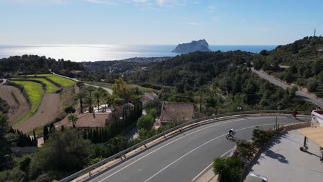 Drone-footage-showing-forested-countryside-and-winding-scenic-hillside-roads-in-Calpe,-Spain,-with-a-cyclist-riding-around-a-corner-in-the-foreground,-and-the-horizon-and-Mediterranean-Sea-behind