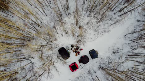 Aerial-Birds-Eye-View-Rise-over-father-and-son-winter-camping-with-summer-and-ice-fishing-tents-forming-a-V-with-a-manmade-campfire-dogs-entertained-on-a-sunset-with-bare-trees-tipped-in-yellow-2-3