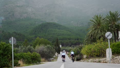 Realtime-low-angle-footage-of-three-team-cyclists-riding-down-a-straight-road-towards-a-forested-hilly-region-in-Calpe,-Spain