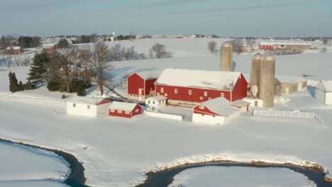 Aerial-reveal-pull-back-shot-of-red-barn,-farm-buildings,-stream-in-rural-USA-during-snow-covered-winter-scene