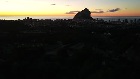 Drone-footage-showing-stunning-silhoette-of-the-town-of-Calpe,-Spain-and-the-natural-landmark-Peñón-de-Ifach,-against-the-backdrop-of-a-beautiful-yellow-and-orange-sunrise-on-an-early-summer-morning