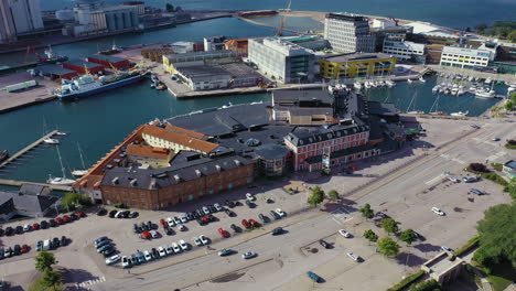 Camera-pans-away-from-shopping-mall-near-seaside-boat-docks-with-large-parking-lot-in-European-city-tourists-attraction-visiting-Denmark-brick-building-aerial-drone-sunny-day-hot-stores-car-vfx-3D