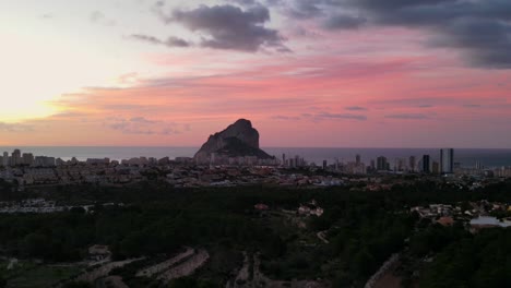 Timelapse-drone-footage-of-beautiful-pink-and-orange-colourful-sunrise-over-town-of-Calpe,-Spain,-showing-the-natural-landmark-Peñón-de-Ifach-and-the-Mediterranean-sea-in-the-background