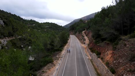 Panning-drone-footage-of-three-team-cyclists-riding-along-a-picturesque-Spanish-road,-through-a-valley-with-exposed-red-earth,-a-river-gorge-and-lush-green-trees-lining-the-road