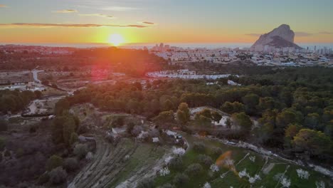 Panning-drone-footage-of-stunning-orange-sunrise-over-Valencian-countryside,-moving-towards-town-of-Calpe,-Spain,-and-the-natural-park-with-landmark-Peñón-de-Ifach