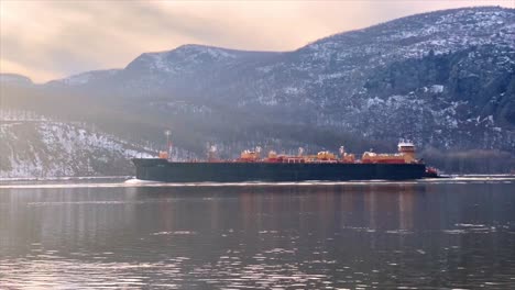 A-cargo-ship-sailing-down-the-hudson-river-in-winter-in-new-york-state-in-the-hudson-valley-during-winter-with-snow-in-the-mountains-and-ice-in-the-water