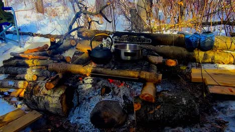 Winter-campsite-fire-log-cabin-style-design-stack-of-wood-with-a-forest-background-snow-covered-grounds-foldable-chair-metal-rack-overtop-the-flames-with-a-tea-kettle-and-a-pot-steaming-boiling-sunset