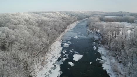 Quick-flying-over-icy-winter-river-surrounded-by-white-treetops-forest