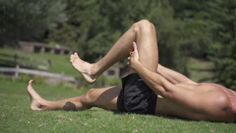 Young-caucasian-fit-attractive-male-exersicing-outdoors-hamstrings-mobility-exercise-with-green-vegetation-background-slow-motion-60-fps