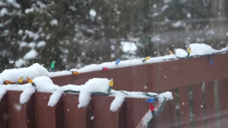 Snow-Falling-Over-Railing-Lined-with-Multi-Colored-Christmas-Lights,-Slow-Motion