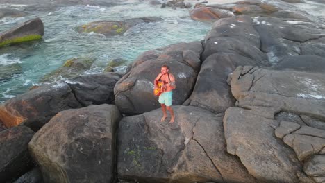 Luftaufnahme-Von-Oben-Nach-Unten-Eines-Jungen-Mannes-In-Badegästen,-Der-Auf-Einem-Felsen-Steht-Und-Gitarre-Spielt,-Vor-Dem-Ozean-Und-Einem-Wunderschönen-Sonnenuntergang,-Australien