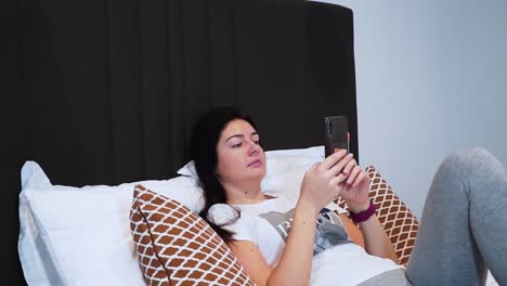 The-young-woman-is-lying-on-the-bed-in-her-bedroom-and-is-scrolling-the-social-media-page-with-her-smartphone,-close-up-shot,-serious-looking