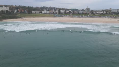 Bondi-Beach-With-Surfers-Riding-The-Waves-With-Eastern-Suburbs-In-Background-At-Sydney,-New-South-Wales,-Australia