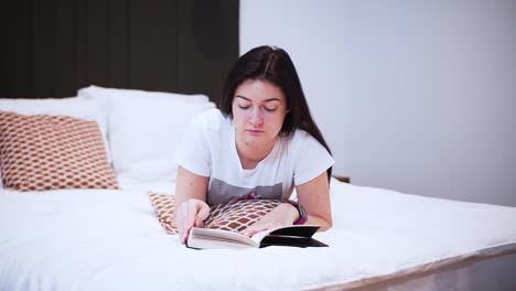 a-young-woman-is-reading-a-book-with-black-hardcover-in-her-bedroom,-serious-looking,-laying-on-the-bed,-she-spends-her-free-time-with-a-book,-relaxing-at-home,-turns-the-page,-flips-the-page