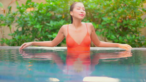 Sexy-asian-woman-in-red-swimsuit-caressing-her-wet-hair-in-a-swimming-pool-with-tropical-plants-in-background,-full-frame-slow-motion