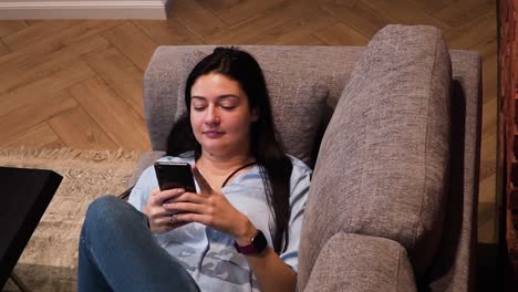 The-young-woman-is-lying-on-the-couch-in-the-living-room-with-a-red-brick-wall-and-is-writing-a-message-with-her-smartphone-and-smiling,-shot-from-the-top,-close-up-shot