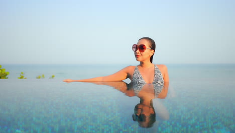 Sexy-happy-asian-woman-in-infinity-swimming-pool-with-sea-horizon-in-background,-mirror-reflection-on-water-on-sunny-day-at-exotic-tropical-location