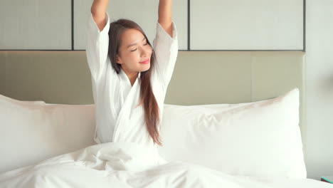 Satisfied-and-smiling-woman-in-bathrobe-stretches-sitting-on-bed