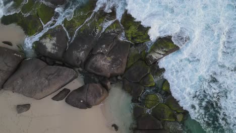 Aerial-Top-Down-View:-Waves-Crash-Against-Rocks-Along-Coastline-and-sandy-beach-during-cloudy-day