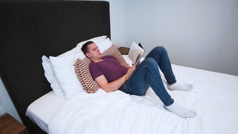 a-young-man-is-reading-a-book-with-black-hardcover-in-her-bedroom,-serious-looking,-laying-on-the-bed,-he-spends-his-free-time-with-a-book,-relaxing-at-home,-turns-the-page,-flips-the-page