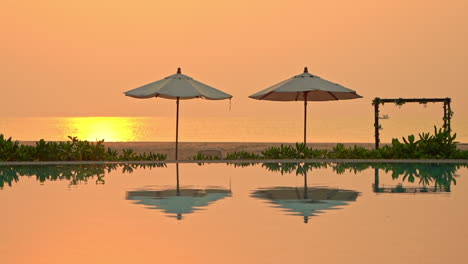 An-orange-sunset-fills-the-evening-sky-reflecting-in-a-swimming-pool-where-two-beach-umbrellas-sit-empty