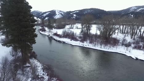 Luftaufnahme---Die-Kamera-Biegt-Sich-Um-Einen-Baum-Und-Enthüllt-Einen-Wunderschönen-Schneebedeckten-Fluss