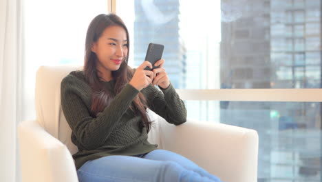 Asiatische-Frau,-Die-Im-Stuhl-Am-Fenster-Sitzt-Und-Mit-Einem-Lächeln-Auf-Dem-Smartphone-Sms-Schreibt