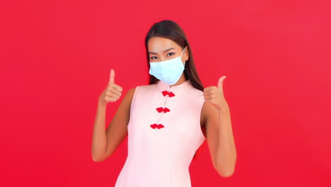 A-young-woman-showing-her-face-mask-and-gives-thumbs-up-hands-gesture,-covid-19-virus-pandemic-concept,-red-background,-static-shot