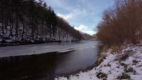 calm-river-with-remaining-ice-and-snow-in-the-woods,-day,-winter,-pan-left