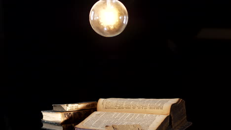 Antike-Bücher-Auf-Dem-Tisch-In-Einem-Dunklen-Raum,-Der-Von-Einer-Glühbirne-Beleuchtet-Wird,-Ein-Gleitender-Dolly-schuss