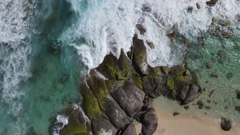 Aerial-top-down-shot-of-crashing-waves-against-rocky-and-mossy-stones-at-shore-of-ocean-with-sandy-beach