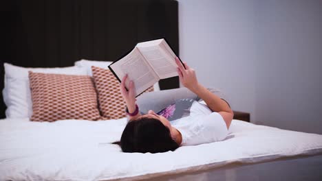 a-young-woman-is-reading-a-book-with-black-hardcover-in-her-bedroom,-laying-on-the-bed,-she-spends-her-free-time-with-a-book,-relaxing-at-home,-turns-the-page,-flips-the-page,-close-up-shot