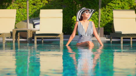 Summer-fashion-concept,-trendy-asian-woman-with-black-and-white-hat-and-swimsuit-sitting-on-swimming-pool-edge-on-sunny-day,-full-frame