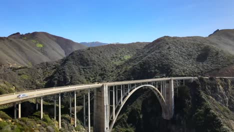 Bixby-Bridge-in-Big-Sur,-aerial-rising-view,-cars-driving-on-bridge,-mountains-on-a-clear-day