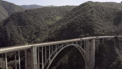 Bixby-Creek-Bridge,-rising-aerial-on-a-clear-day,-with-cars-traveling-across,-Big-Sur