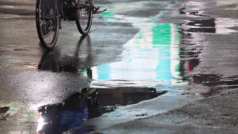 Reflection-Of-Person-On-Bicycle-Seen-At-Puddle-On-Asphalt-Road-After-Raining-At-Night-In-Tokyo,-Japan