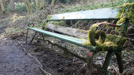 Mossy-overgrown-old-weathered-green-curled-wrought-iron-wooden-seat-abandoned-in-woodland