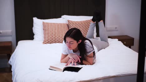 a-young-woman-is-reading-a-book-with-black-hardcover-in-her-bedroom,-laying-on-the-bed,-she-spends-her-free-time-with-a-book,-relaxing-at-home,-turns-the-page,-flips-the-page,-close-up-shot,-smiling