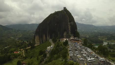 Drone-Flies-Away-From-La-Piedra-del-Penol,-with-Crowded-Parking-Lot-Full-of-Tourists
